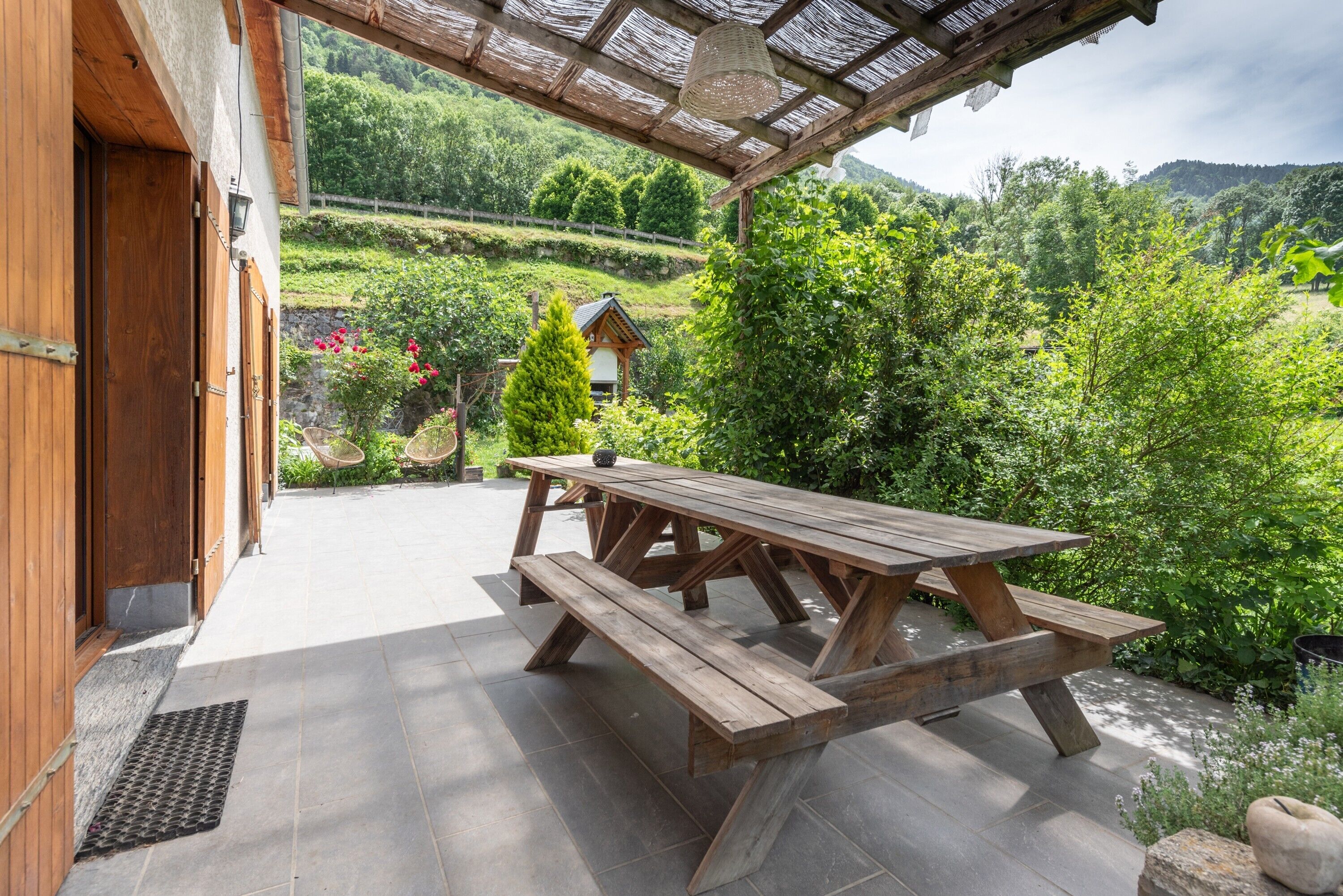 Een houten picknicktafel met banken, een pergola en uitzicht op een weelderige tuin en verre heuvels.
