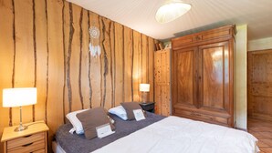 A bedroom with a wooden wall, a bed with white and brown bedding, two bedside tables with lamps, and a wardrobe.