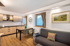 A modern kitchen with wooden cabinets, a dining table with chairs, a sofa with a patterned pillow, a clock, and a framed landscape painting on the wall.