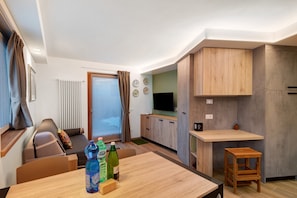 A compact living space with a sofa, a dining table, a kitchen area with cabinets, a television, and a small wooden stool.