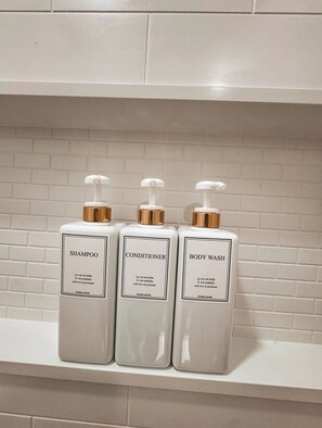 Three bottles of HARRA HOME shampoo, conditioner, and body wash on a white shelf against a white brick wall. - Charming 2-bedroom house with WiFi, AC in Spartanburg (Spartanburg)