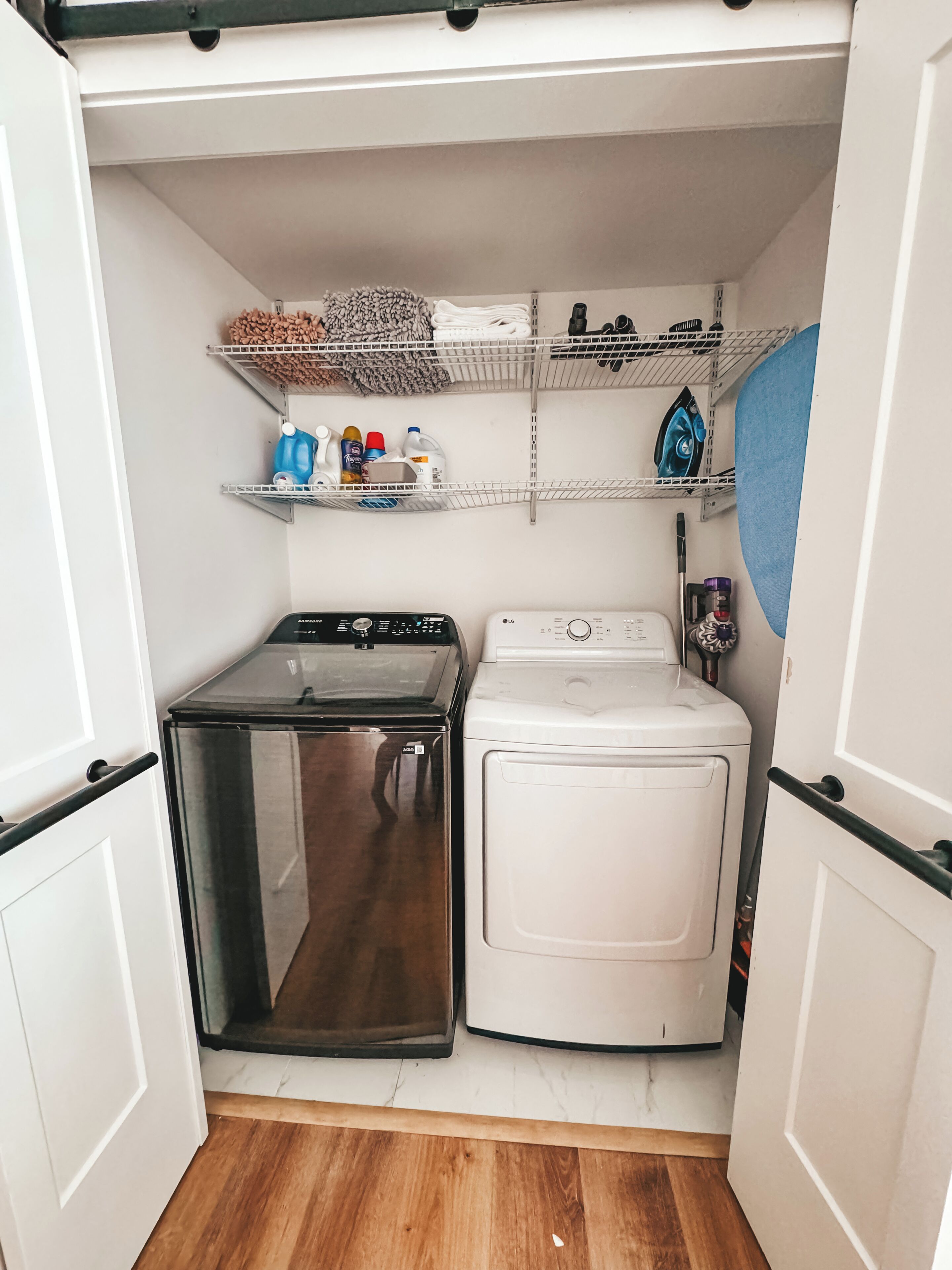 A compact laundry room with a stainless steel dryer, a white washing machine, and shelves above holding cleaning supplies.