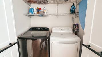 A compact laundry room with a stainless steel dryer, a white washing machine, and shelves above holding cleaning supplies.