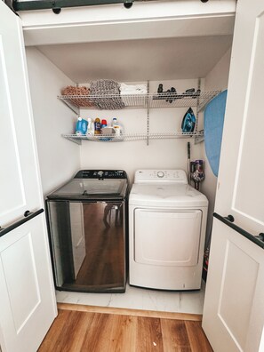 Un lavadero pequeño con una secadora de acero inoxidable, una lavadora blanca y estantes superiores con productos de limpieza.