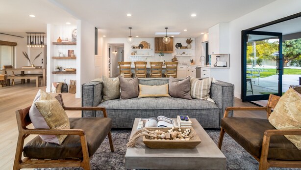 Ein modernes Wohnzimmer mit einem grauen Sofa, Holzstühlen und einem Couchtisch mit Büchern und einer dekorativen Schale.