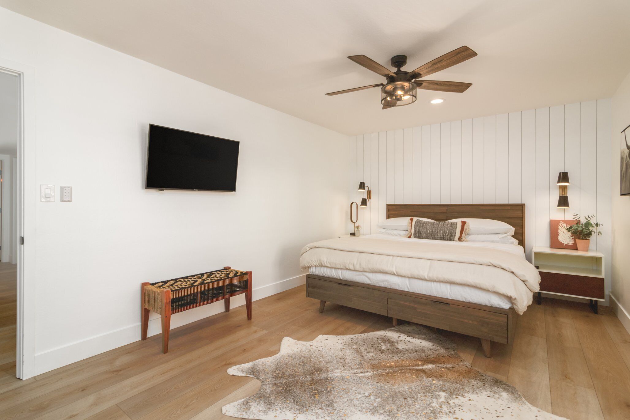 A bedroom with a wooden bed, a ceiling fan, a mounted TV, a bench, and a rug.