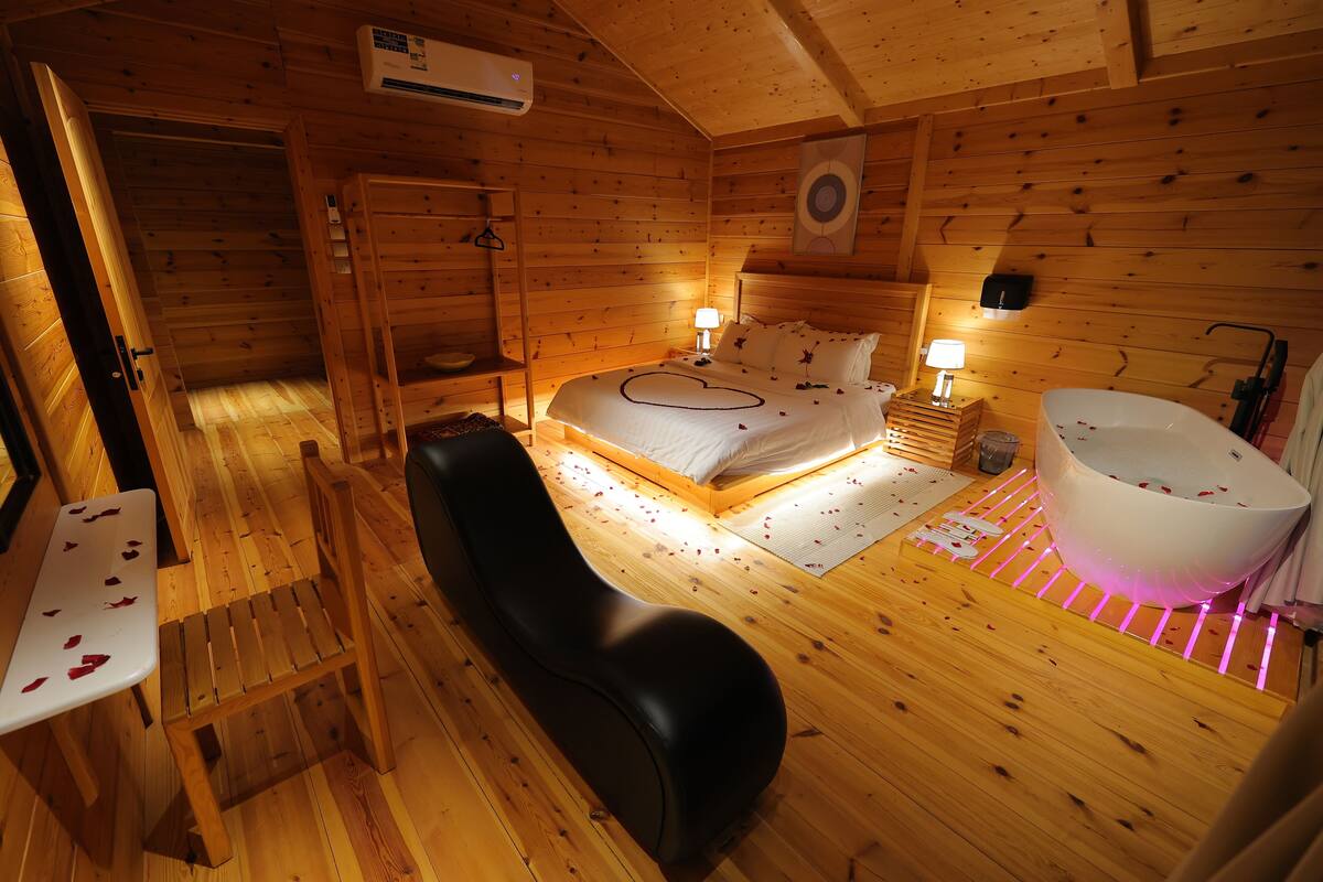 A wooden cabin bedroom with a bed, bedside lamps, a bench, a sink, and a wooden floor.