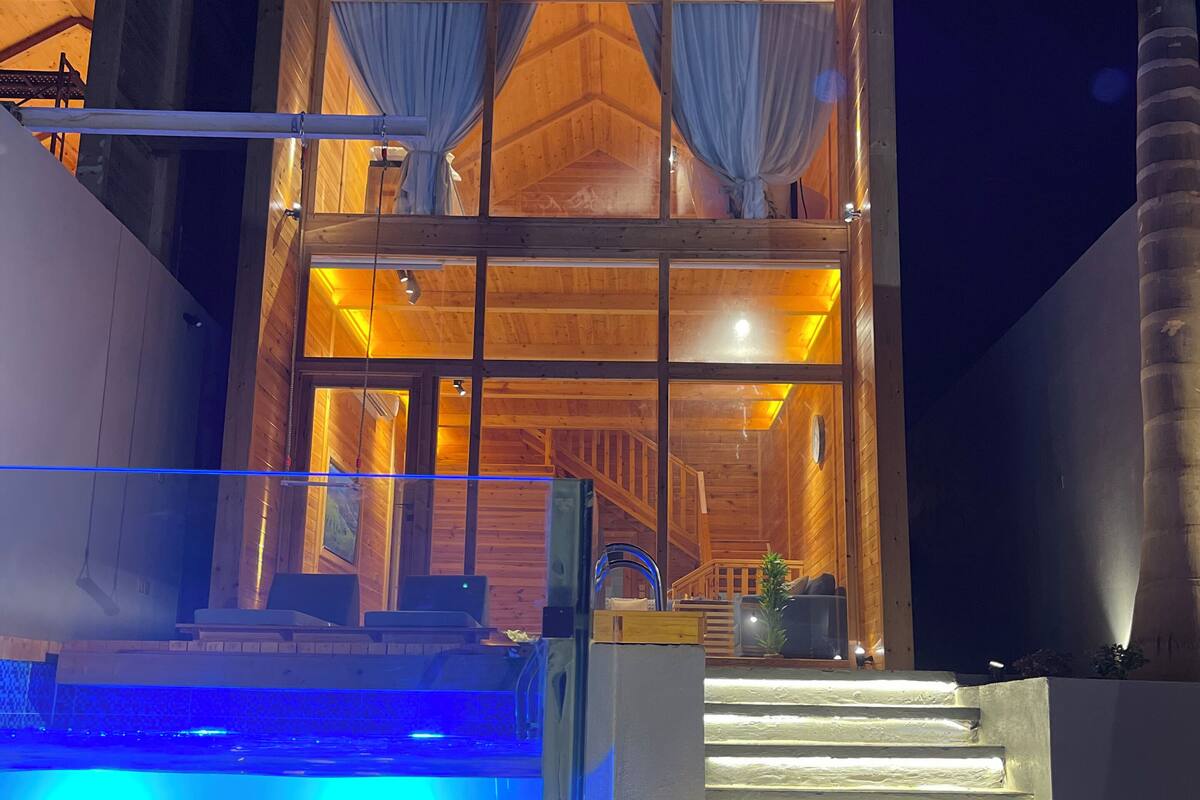 A modern wooden building with a large glass facade, a visible staircase leading to the entrance, and a pool area with blue lighting.