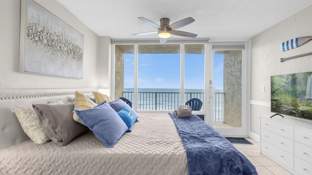 Une chambre avec un lit, un ventilateur de plafond, une grande fenêtre et une vue sur l’océan.