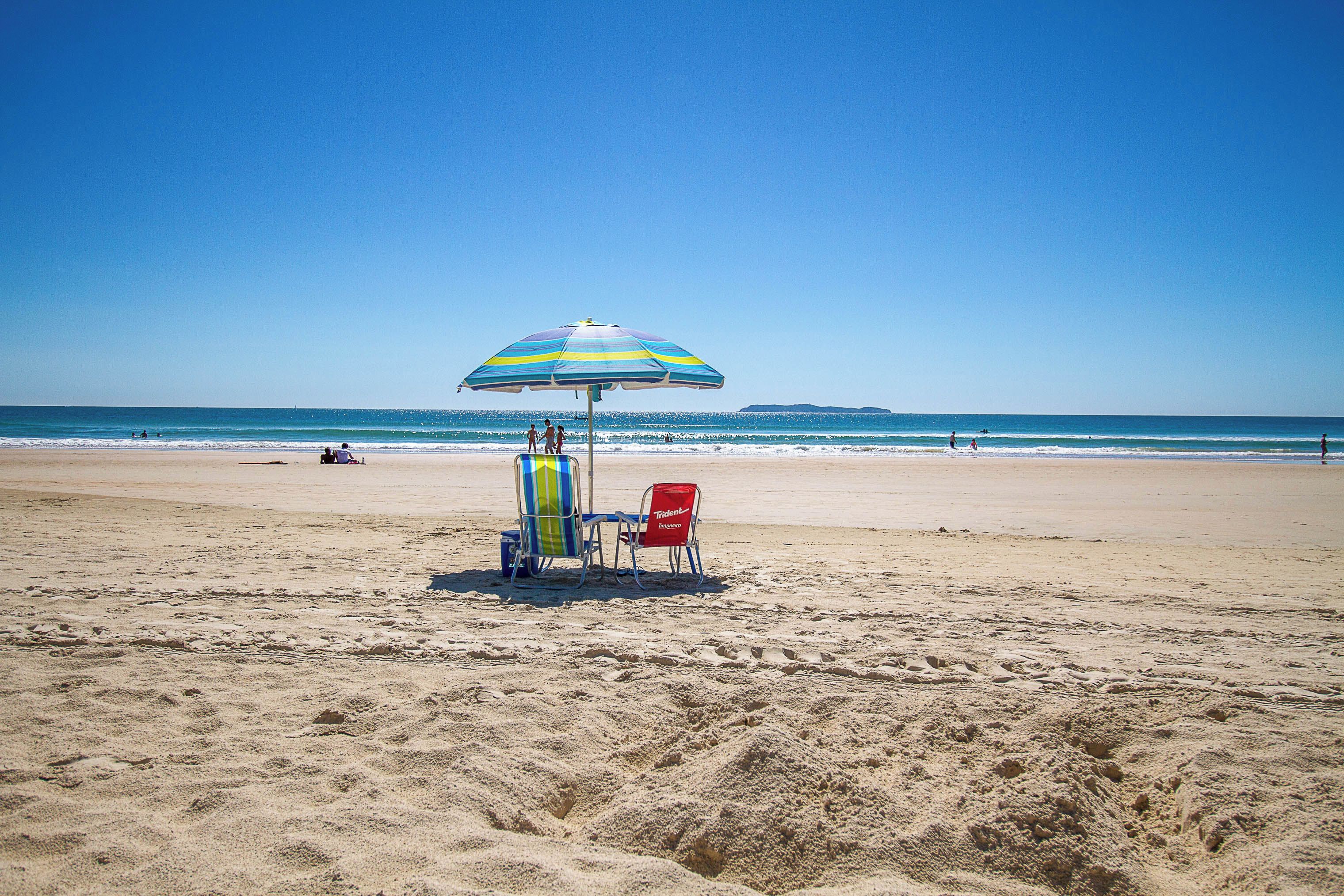A beach with a striped umbrella, two beach chairs, and a cooler on the sand.