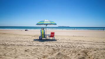 A beach with a striped umbrella, two beach chairs, and a cooler on the sand.