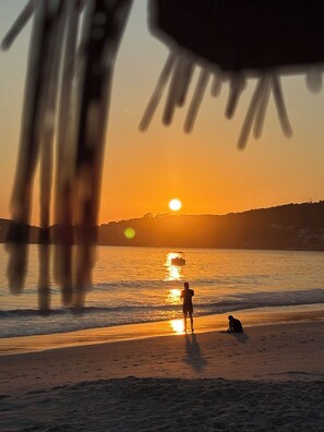 A beach scene with the sun setting over the water, silhouettes of people on the sand, and a distant shoreline. - Loft for a House in Bombinhas - NO GARAGE (Centro Bombinhas)