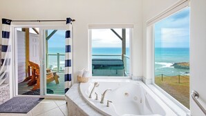 A bathroom with a large bathtub, a glass door leading to a balcony with a view of the ocean, and a wooden deck with outdoor furniture. - Starfish by OceanviewHotTubs Oceanfront Tesla charging  Shelter Cove (Whitethorn)