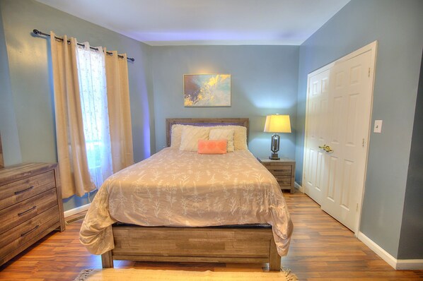 A bedroom with a bed, a nightstand, a lamp, a dresser, a door, and a window with curtains.