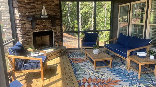 A cozy outdoor seating area with a stone fireplace, wooden furniture, and large windows offering a view of the forest. - Cozy Cabin in Lake Community (Tallassee)