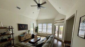 A spacious living room with a large sectional sofa, a wooden coffee table, and a ceiling fan. There are windows with blinds, a bookshelf, and a decorative sailboat model. - Cozy Cabin in Lake Community (Tallassee)