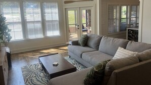 A living room with a sofa, coffee table, and a view of the backyard through large windows. - Cozy Cabin in Lake Community (Tallassee)
