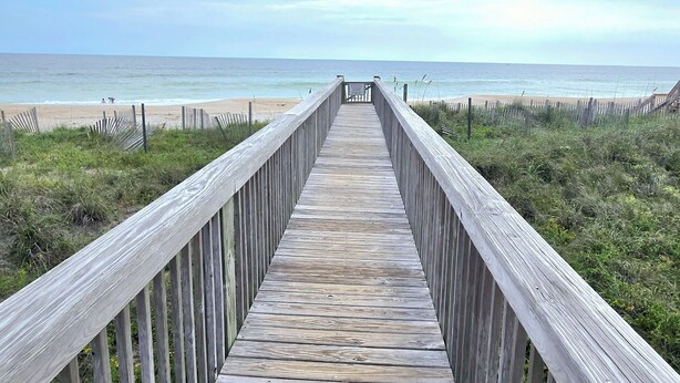 Ein hölzerner Steg führt zu einem Strand unter blauem Himmel.