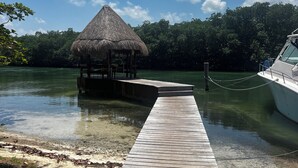 Une jetée en bois mène à une construction à toit de chaume, située au bord d’une étendue d’eau calme, et entourée d’une végétation luxuriante.
