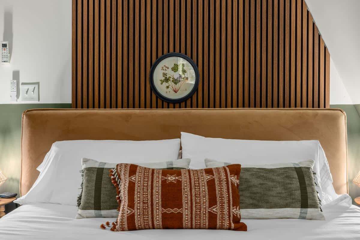 Un dormitorio con cabecera de listones de madera, un grabado botánico enmarco y dos almohadas estampadas.