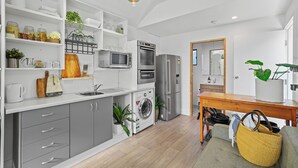 Une cuisine moderne avec des meubles blancs, un four encastré, un micro-ondes et un réfrigérateur. On y trouve une table à manger en bois avec des chaises, ainsi qu’un lave-linge dans un coin.