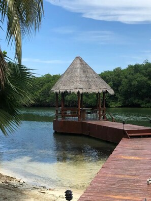 A wooden gazebo with a thatched roof on a dock extending into a calm body of water, surrounded by lush greenery. - HERMOSA CASA FRENTE A LA LAGUNA (Cancún)