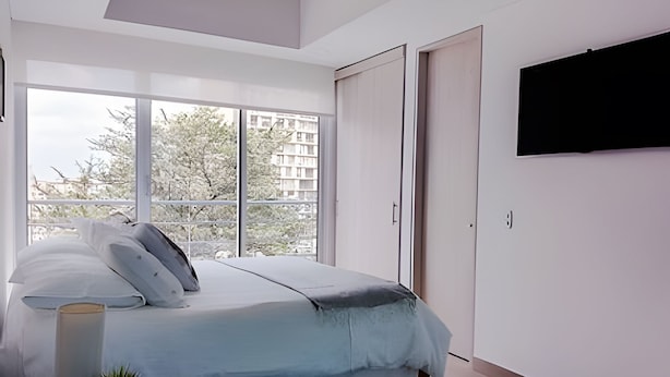 Quarto com cama, televisão fixada na parede e janela com vista para edifícios e árvores.
