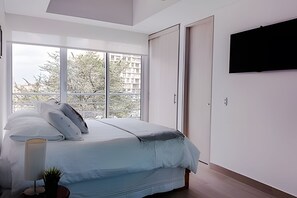 Un dormitorio con cama, televisor montado en la pared y ventana con vistas a edificios y árboles.
