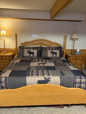 Ein Schlafzimmer mit einem Holzkopfteil, einer gesteppten Bettdecke mit Elchmotiven, zwei Nachttischen inklusive Lampen und einer hölzernen Bank am Fußende des Bettes.