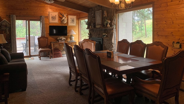 A wooden cabin interior with a dining table, chairs, a fireplace, a television, and a ceiling fan.
