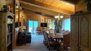 A cozy cabin interior with a wooden ceiling, a dining area with a large table and chairs, a fireplace, and a television mounted on the wall.
