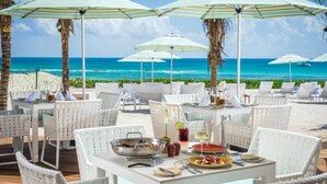 En restaurang vid stranden med vita flätade stolar och bord, parasoller och utsikt över havet.