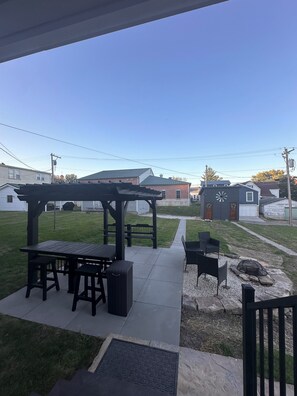 A patio area with a pergola, tables, and chairs, a fire pit, and a stone pathway leading to a house with a clock and wreaths.