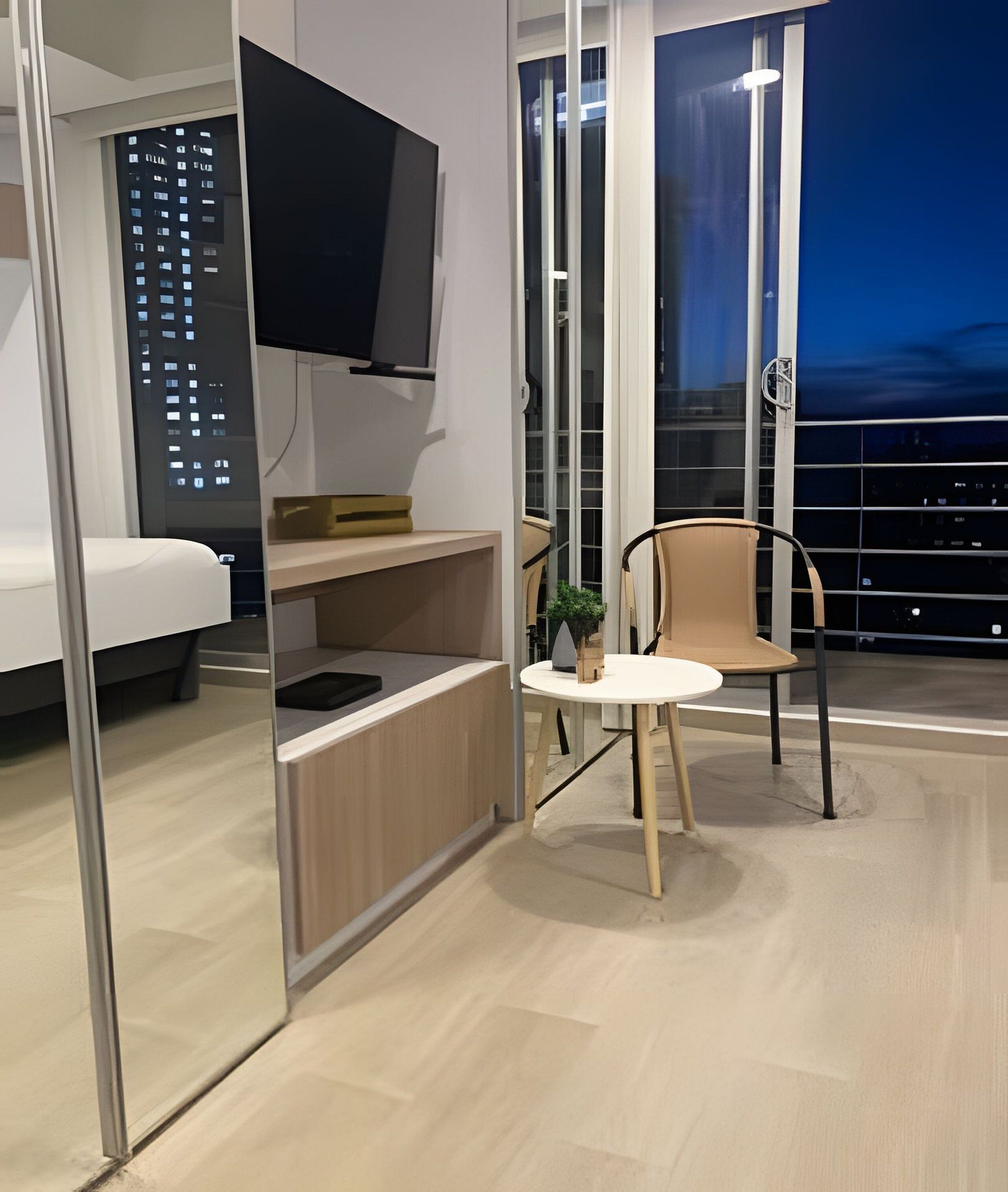 A modern hotel room with a bed, a small kitchenette, a TV, a balcony with a chair, and a view of the city at night.