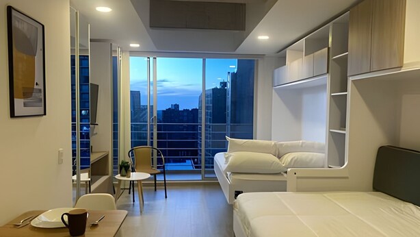 A compact hotel room with a balcony, a bed, a small table with a cup, and a chair. - Maravilloso loft con vista 707 (Bogotá)