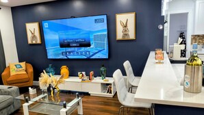 A modern living room with a flat-screen TV, two framed rabbit pictures, a glass coffee table, a sofa, and a dining area with chairs and a bar. - Miramar Game Room Home Arcade · King Bed  15 Min to Beach (Miramar)