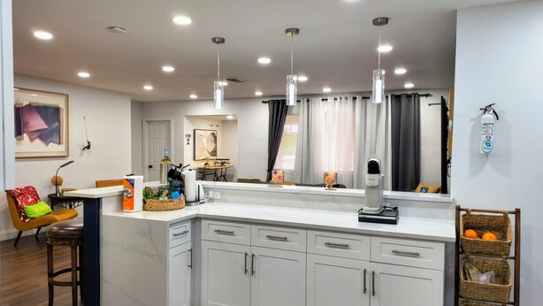 A modern kitchen with white cabinets, a marble countertop, pendant lighting, and a dining area with a chair and table. - Miramar Game Room Home Arcade · King Bed  15 Min to Beach (Miramar)