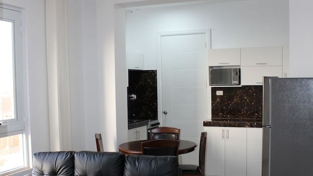 A modern living room with a black leather sofa, a glass coffee table, and a dining area with a wooden table and chairs. The kitchen features white cabinets, a microwave, and a refrigerator. - Blanca Rosa's Place (Pacasmayo)