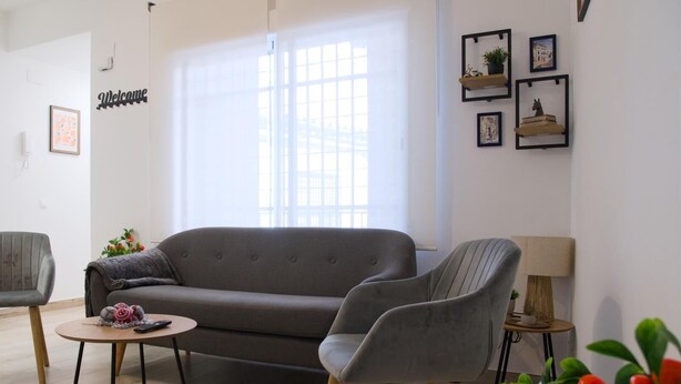 Ein Wohnzimmer mit grauem Sofa, zwei Sesseln, einem Couchtisch und einem kleinen Beistelltisch mit Lampe. An der Wand hängt ein Schild mit der Aufschrift 'Welcome', ein Fenster mit Jalousien und einige gerahmte Bilder sind zu sehen.