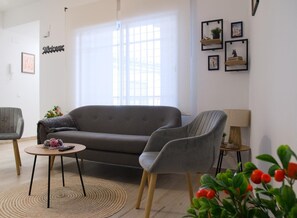 A living room with a grey sofa, two armchairs, a coffee table, and a small side table with a lamp. There is a 'Welcome' sign on the wall, a window with blinds, and a few framed pictures. - CAROL HOME CORDOBA APT (Córdoba)