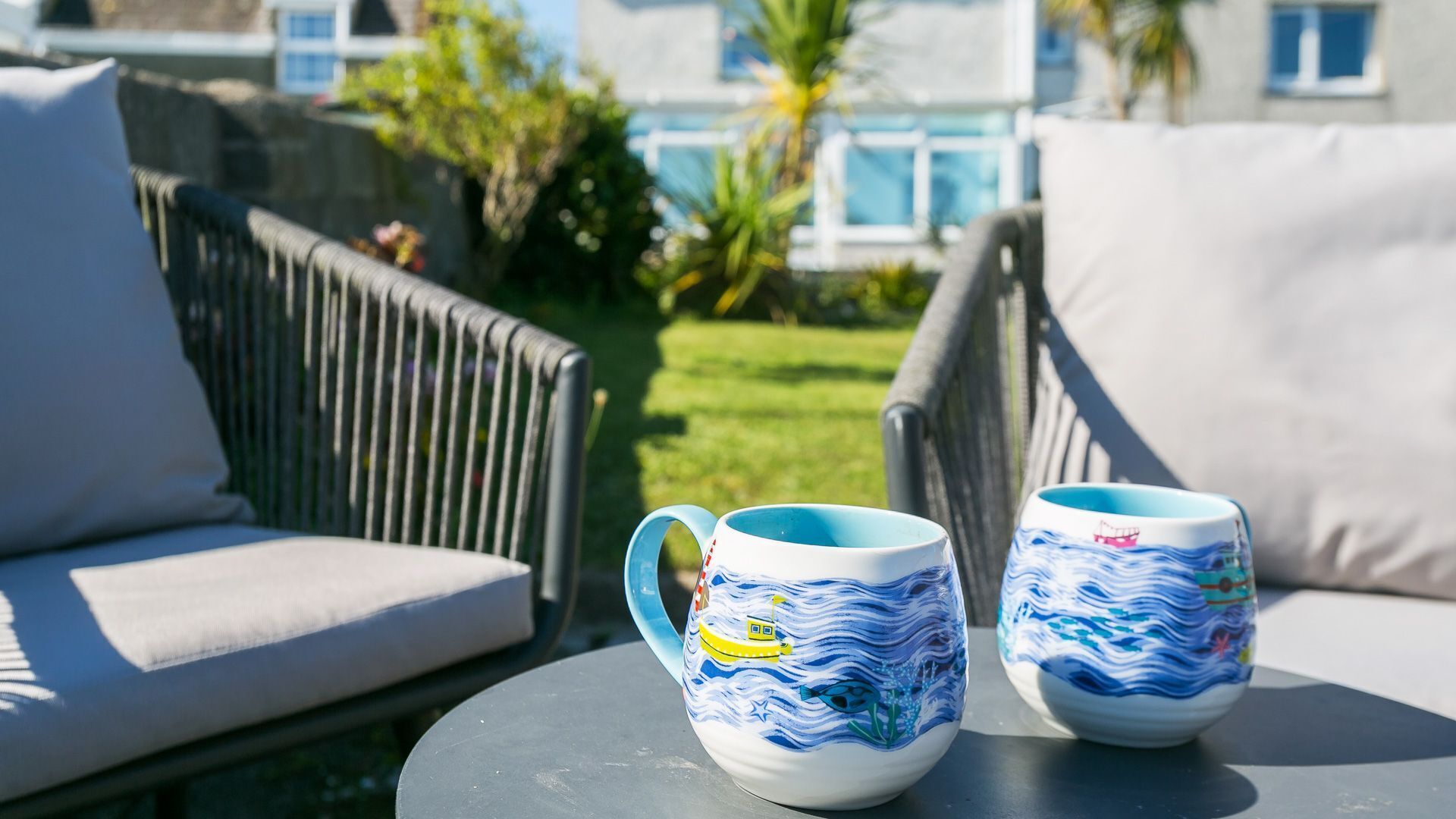 Deux tasses en céramique décorées sur le thème de la marine, posées sur une table ronde dans un espace patio aménagé avec des sièges d’extérieur.