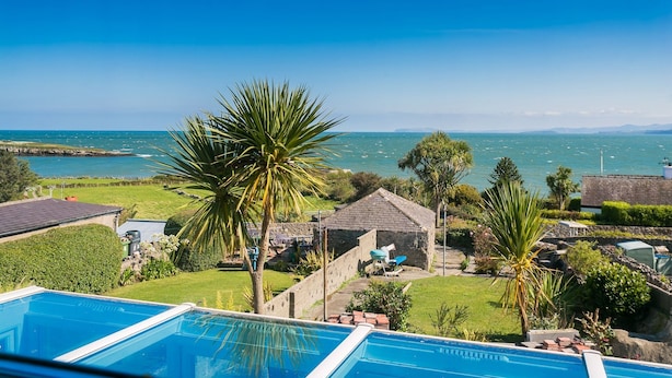 A pool area with clear views of the sea and boats, surrounded by lush greenery and a stone building. - 7 Seaview - sleeps 4 guests  in 2 bedrooms (Moelfre)