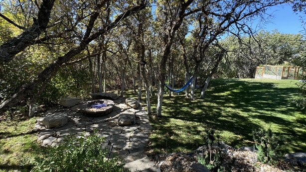 Un foyer en pierre entouré de rochers et de verdure, un hamac et une pergola en bois dans un jardin à l’arrière d’une maison, bordé d’arbres.