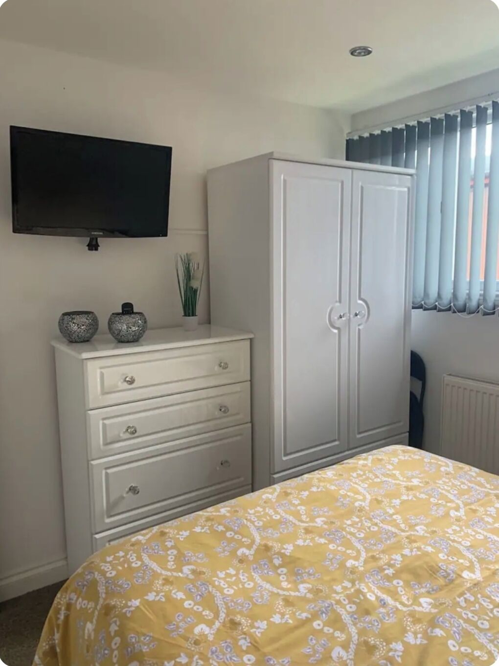 A bedroom with a bed, a white dresser with drawers, a television on the wall, and a window with curtains.