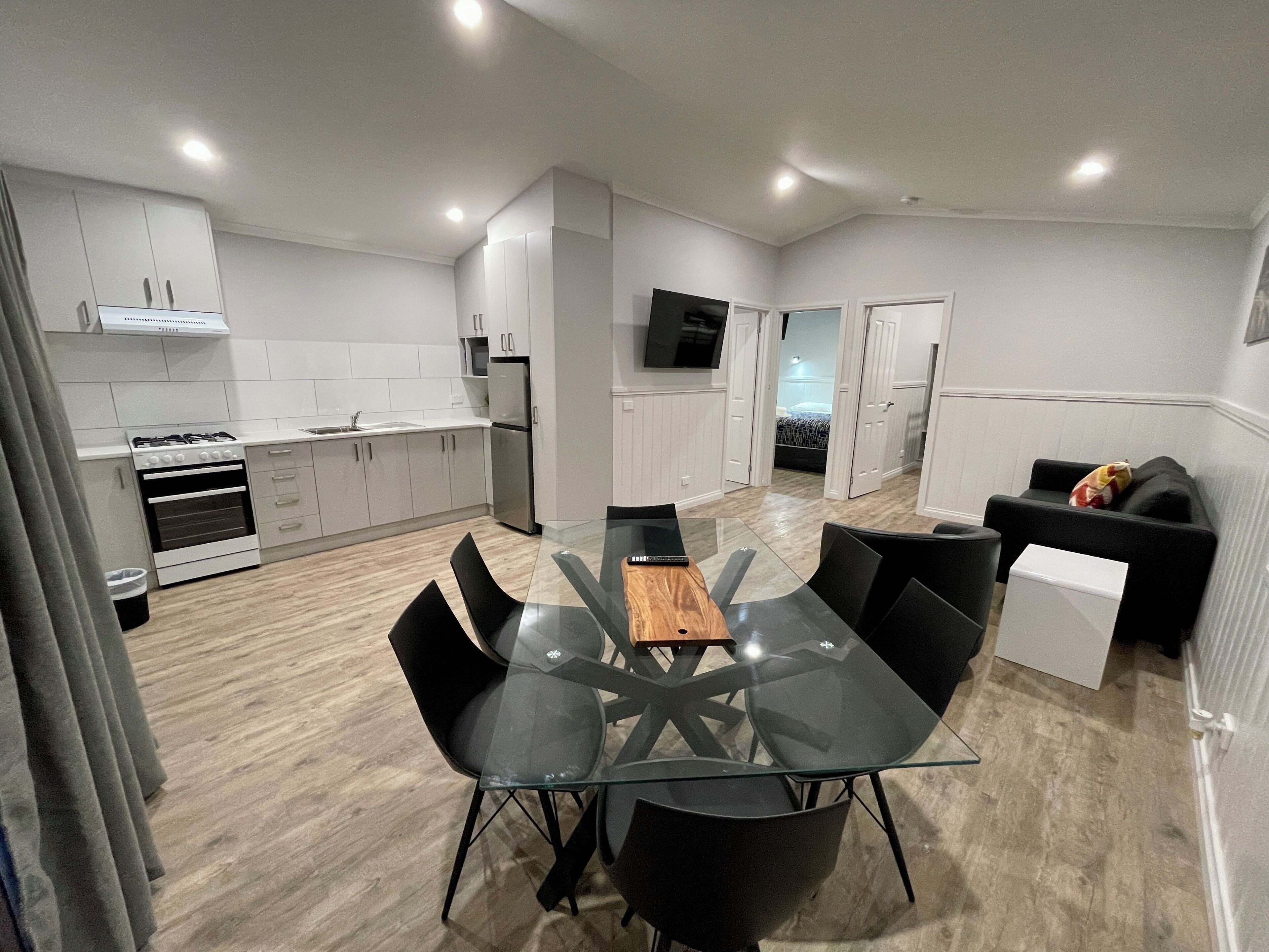 Una cocina moderna con armarios blancos, una estufa y un refrigerador. Hay un comedor con una mesa de vidrio y sillas negras. En la sala de estar hay un sofá negro.