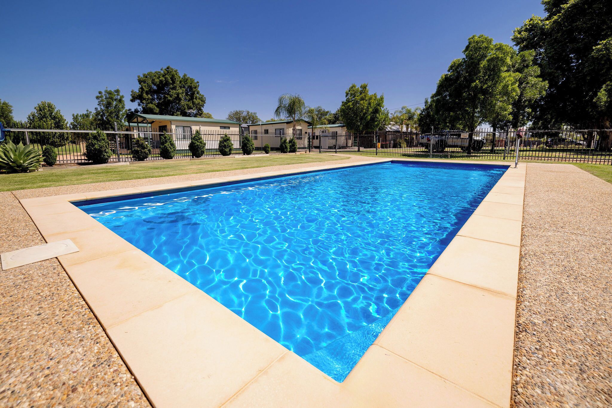 A rectangular swimming pool with clear blue water, surrounded by a paved deck and a metal fence.