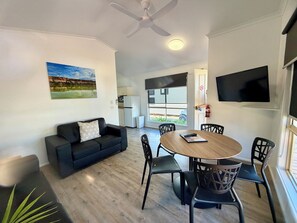 A modern living room with a black sofa, a wooden dining table with chairs, a ceiling fan, a flat-screen TV mounted on the wall, and a large window with blinds. - BIG4 Red Cliffs Holiday Park (Red Cliffs)