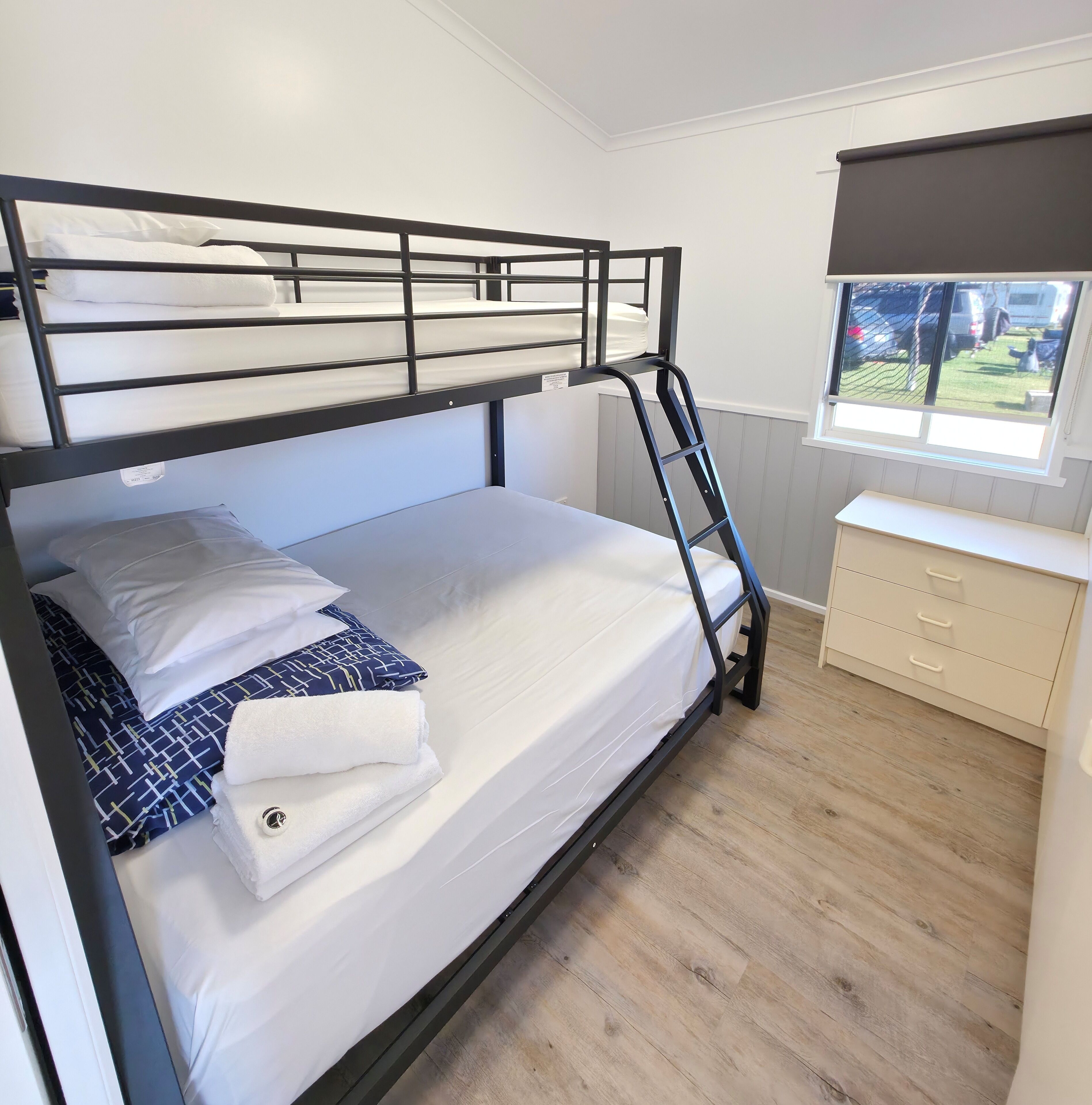 Una cama litera con ropa de cama blanca y una escalera, un aparador con cajones, y una ventana con vistas al exterior.
