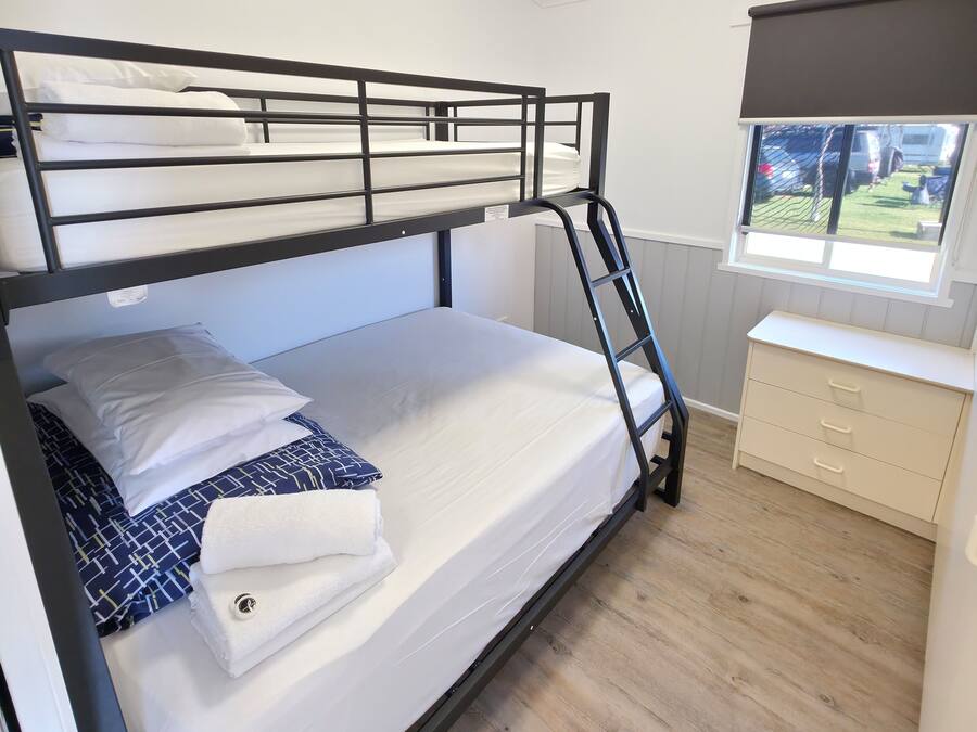 Una cama litera con ropa de cama blanca y una escalera, un aparador con cajones, y una ventana con vistas al exterior.
