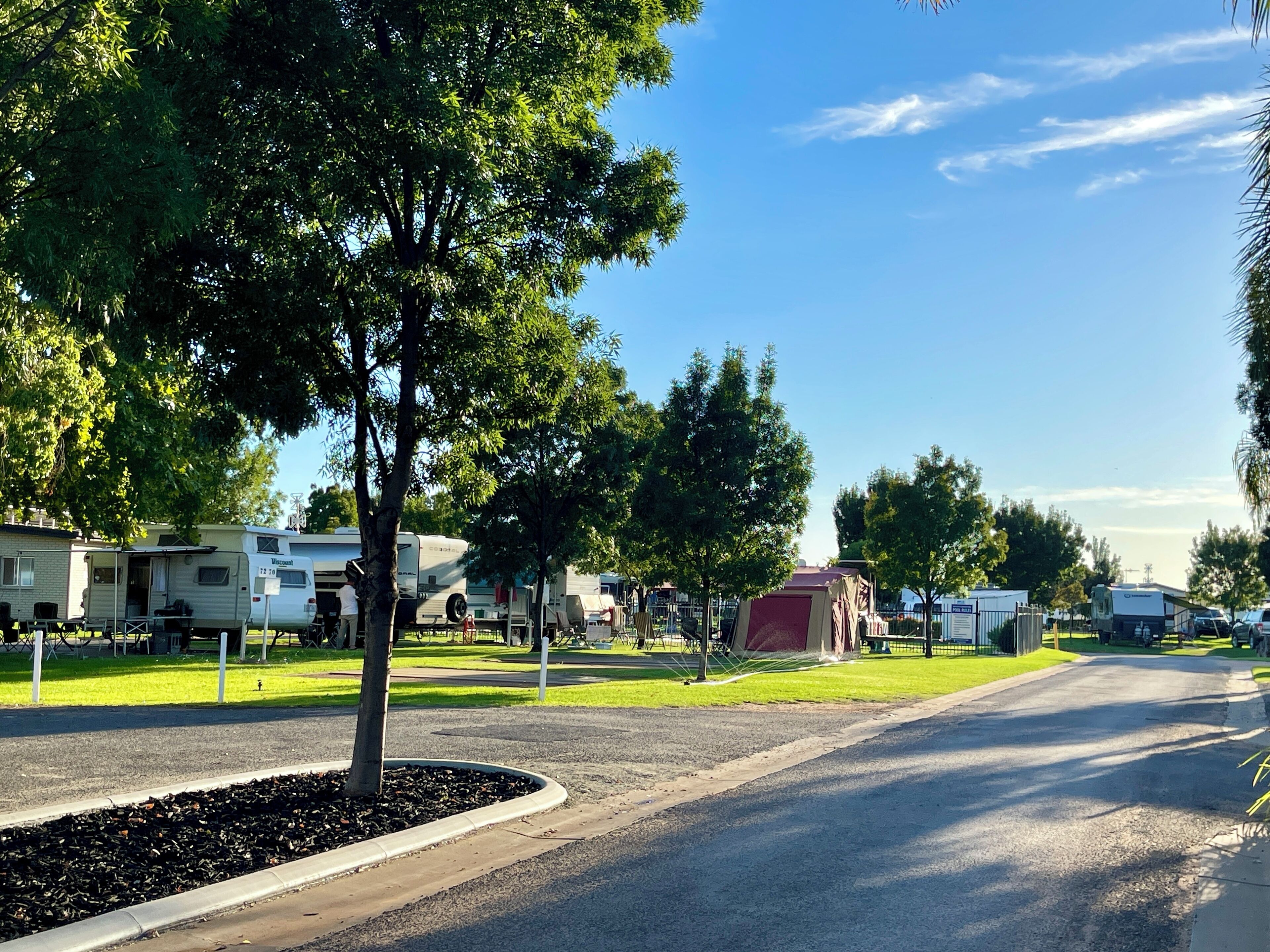 Un camping con vehículos recreativos estacionados en una calle pavimentada, rodeado de árboles y áreas verdes.